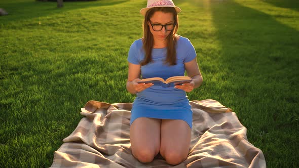Girl in Glasses Reading Book Sitting on a Blanket in the Park at Sunset alt