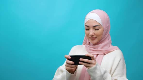 Young Muslim Woman in Hijab Eagerly Playing Video Game on Mobile Phone Enjoying Gameplay in Shooter