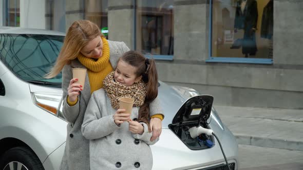 A Woman with Her Daughter Drink Coffee Near Her Electric Car. Charging an Electric Car at a Gas alt