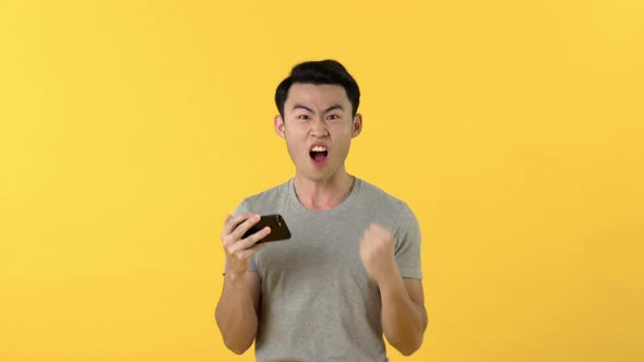Excited Asian man looking at smartphone and cheering with fist pumping gesture