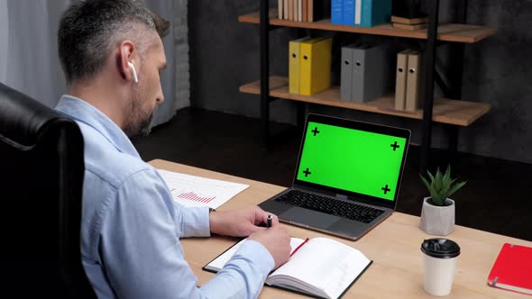 Man Stock Trader Broker Makes Notes in Notebook Uses Laptop with Green Screen alt