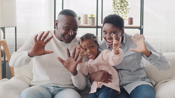 Afro American Happy Family Sitting at Home Sofa Waving Hello Looking at Camera Little Girl Daughter alt