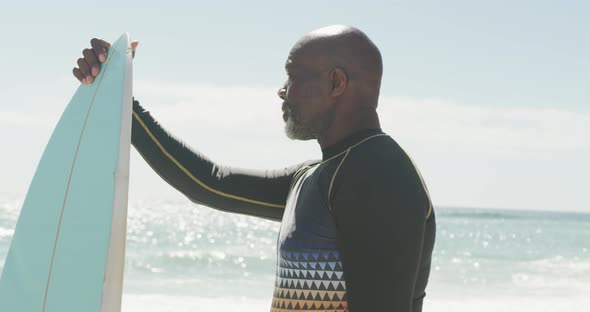 Senior african american man standing with surfboard on sunny beach alt