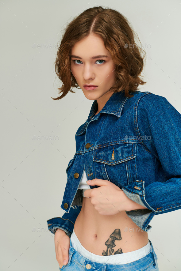 attractive young model with tattoo in cropped denim jacket and blue jeans  posing on grey backdrop Stock Photo by LightFieldStudios