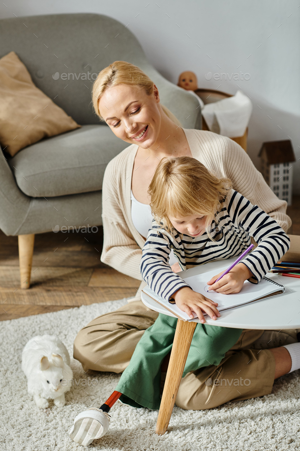 blonde girl with prosthetic leg drawing on paper with colorful pencils near happy mother at home