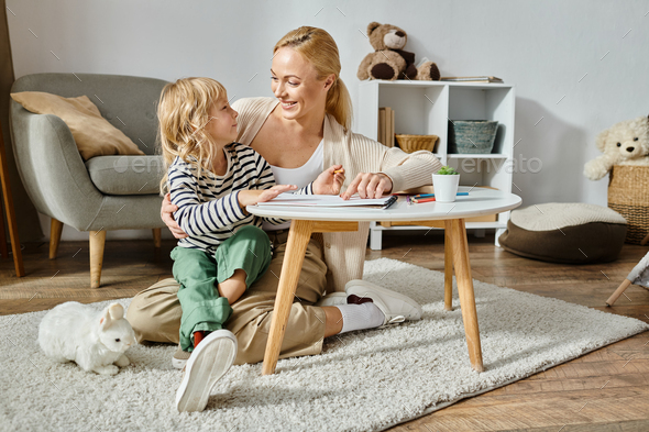 happy blonde woman looking at her daughter with prosthetic leg drawing on paper with colorful pencil