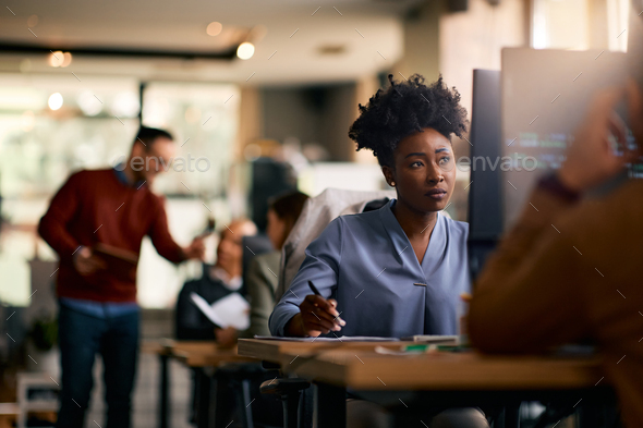 Black female computer programmer taking notes while working on desktop PC in the office.