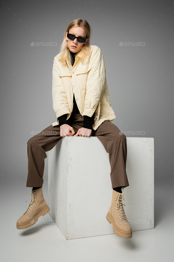 vertical shot of young androgynous person in stylish attire posing on huge cube in trendy sunglasses