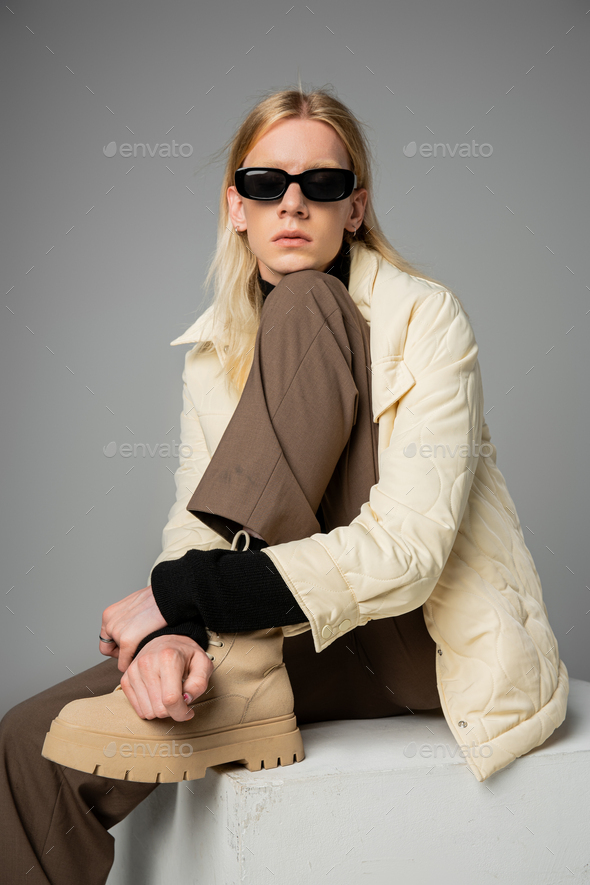 good looking androgynous person in trendy winter attire sitting on huge white cube, fashion concept