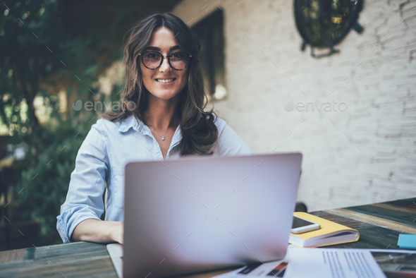 Joyful adult woman looking at camera while using laptop on terrace
