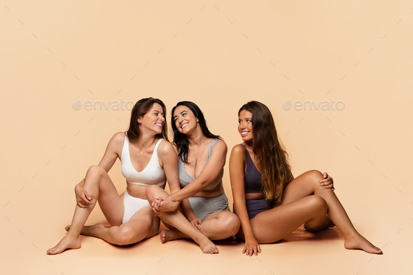 Three glad diverse millennial women in undergarments sit on floor