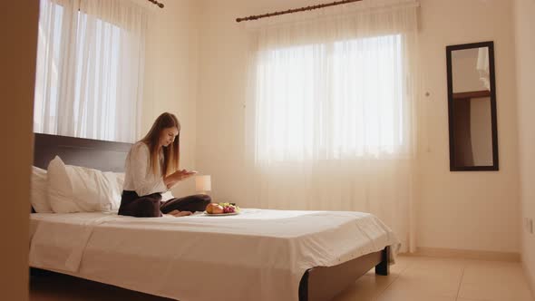 Happy Young Woman Sitting on the Bed with a Plate of Fresh Fruits and Taking alt