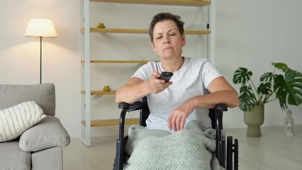 Senior Lady in Wheelchair Watching Tv in Living Room Holding Remote Control Changing Tv Channels alt