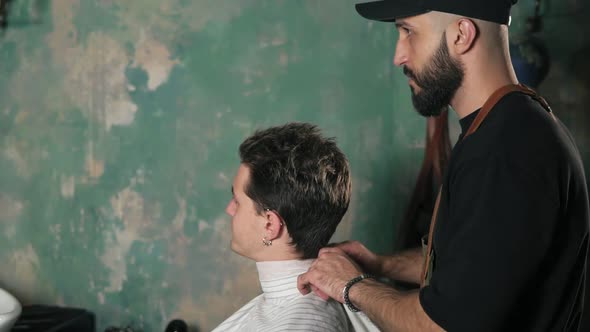 Attractive Young Man Sitting in a Chair While Bearded Barber is Preparing Him for a Haircut alt
