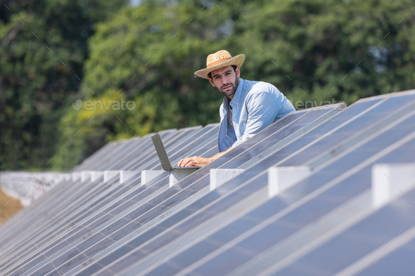 young male technician in sun hat and shirt holding laptop and checking solar panels
