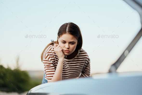 A woman with a wrench looks sadly and sadly under the open hood of her car and can\'t fix it from a