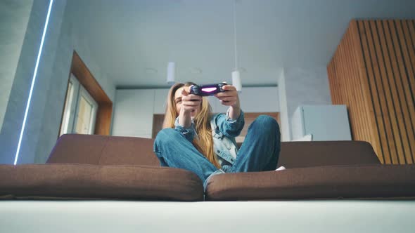 A Beautiful Excited Young Gamer Girl is Sitting on a Couch and Playing Video Games on a Console alt