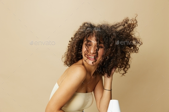Woman dries curly afro hair with blow dryer, home beauty care styling products hair, smile on beige