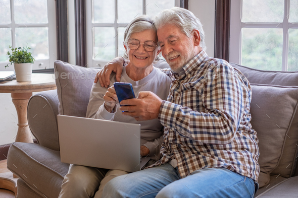 Carefree 70s white-haired senior couple sitting on couch in living room using phone and laptop