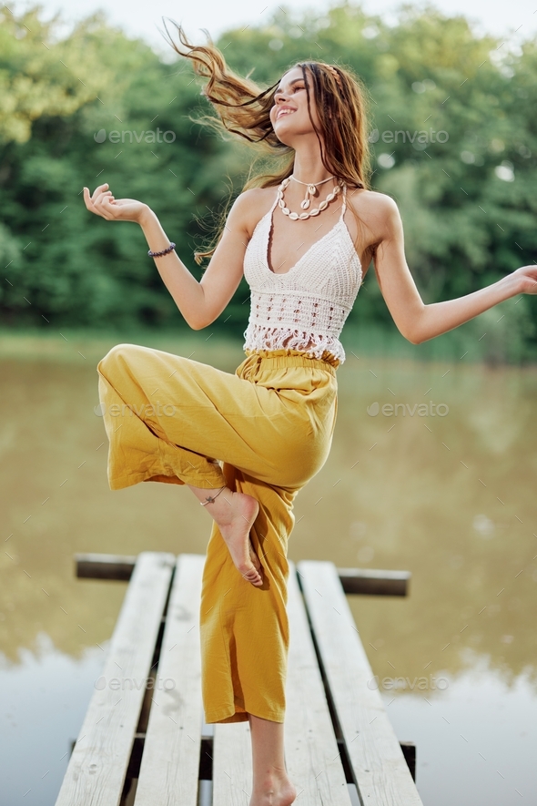 A young woman in a hippie look and eco-dress dancing outdoors by the lake wearing a hat and yellow