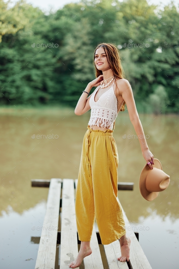 A young woman in a hippie look and eco-dress dancing outdoors by the lake wearing a hat and yellow