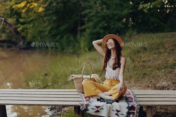 Woman in hippie eco dress sits in nature by the lake on a bridge relaxing with a basket of food and