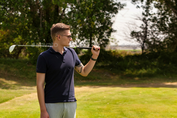 Serious young guy golfer in sunglasses with golf club on shoulder training in golf course