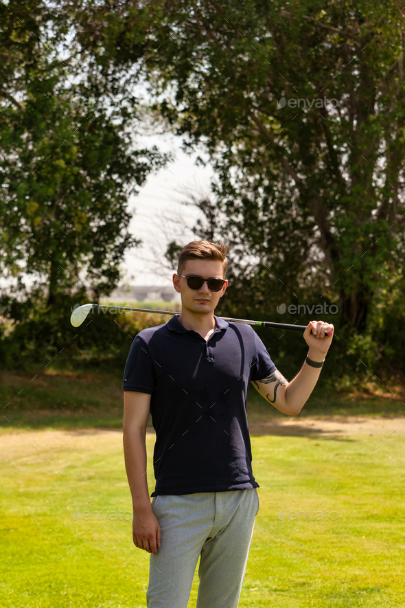 Confident young guy golfer in sunglasses with golf club on shoulder posing in golf course