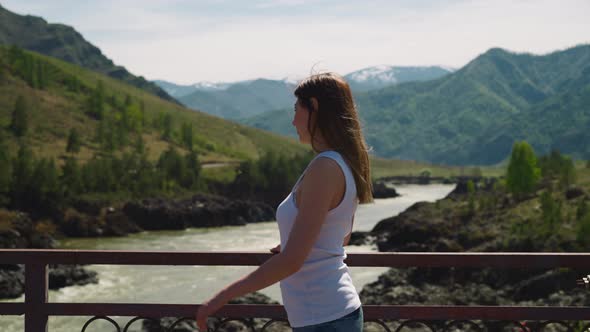 Woman Looks Around on Bridge Over River in Mountain Valley alt