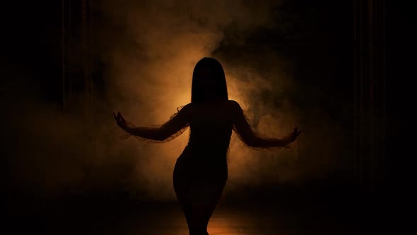 Dark Silhouette of Young Woman Dancing in Dark Smoky Studio Against Background of Yellow Lights alt