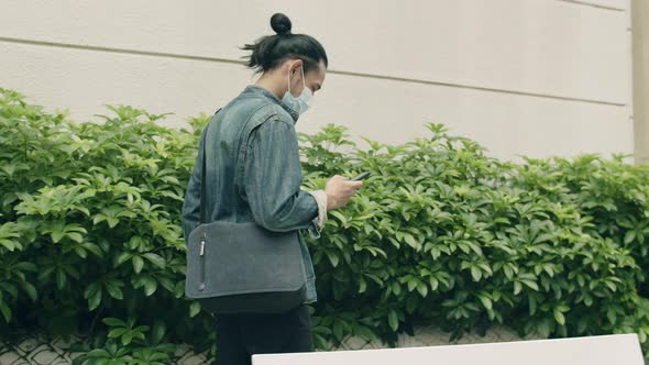 Asian man wearing a surgical mask and using a smartphone while walking on the street.