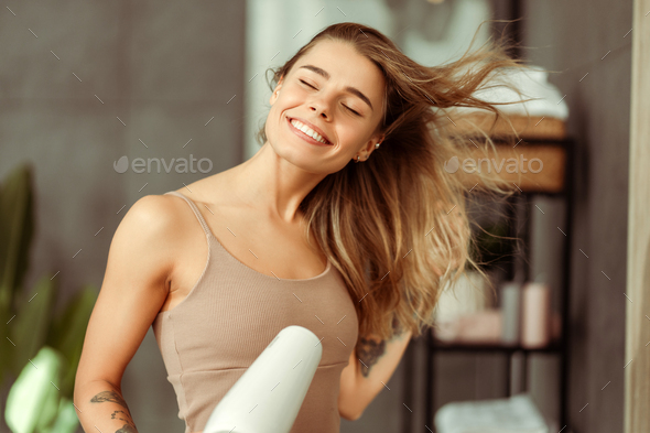 Happy beautiful young woman drying her hair with dryer, wearing tank top standing in bathroom