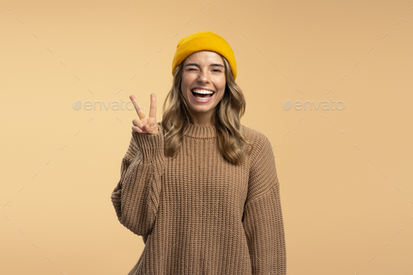 Smiling beautiful woman wearing stylish yellow hat and winter brown sweater  showing victory sign Stock Photo by msvyatkovska