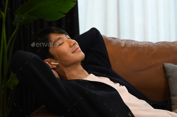 Calm happy asian man lying on comfortable couch enjoy stress free peaceful mood at home.