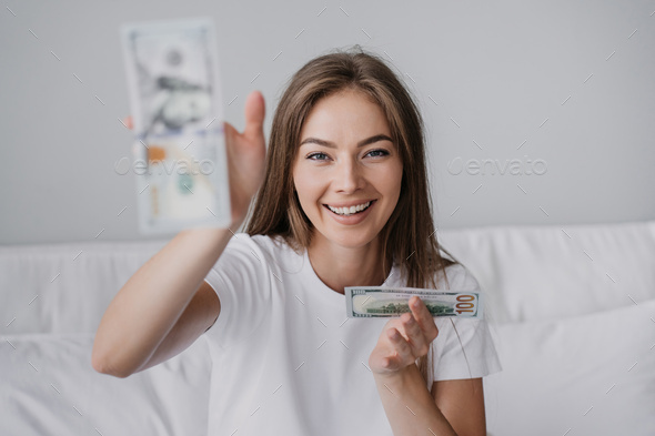 Beautiful Italian girl sitting on bed throwing money dollar banknotes at camera toothy smiles.