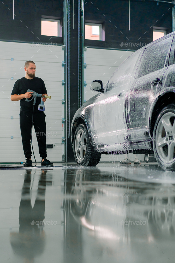 A male car wash employee applies car wash foam to a luxury black car using spray gun in car wash box