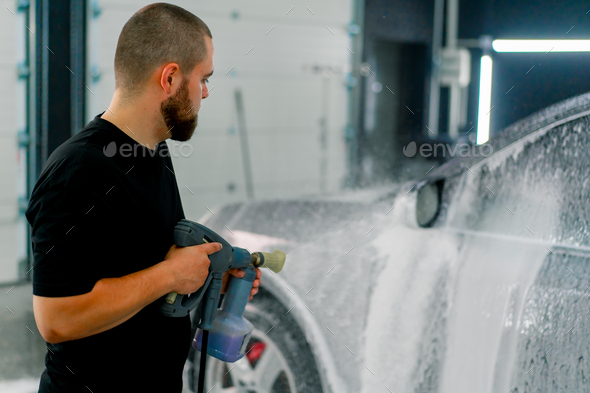 A male car wash employee applies car wash foam to a luxury black car using spray gun in car wash box