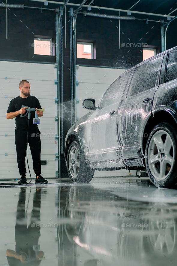 A male car wash employee applies car wash foam to luxury black car using spray gun in car wash box