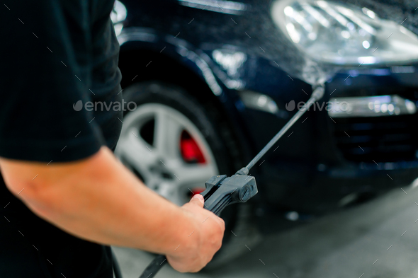A male car wash employee applies car wash detergent to a black car using spray gun in car wash box