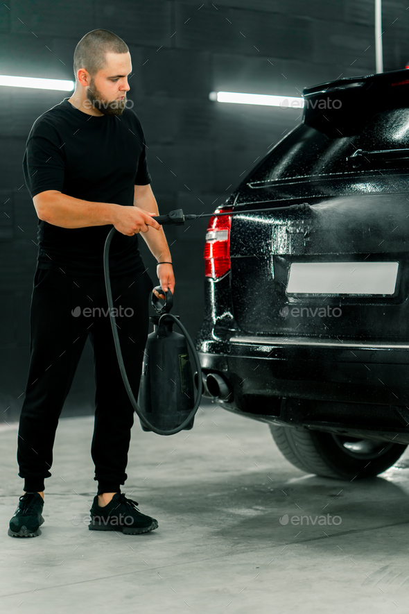 A male car wash employee applies car wash detergent to a black car using spray gun in car wash box