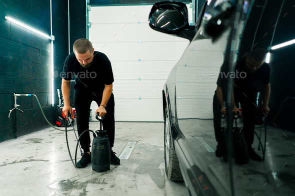 A male car wash employee applies car wash detergent to black car using spray gun in car wash box