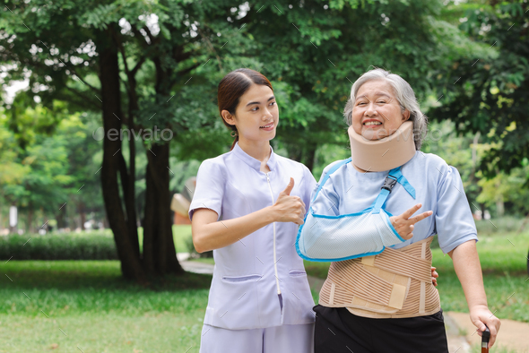 Senior Asian woman wearing patient gown and soft splint due to sore arm showing okay sign with nurse