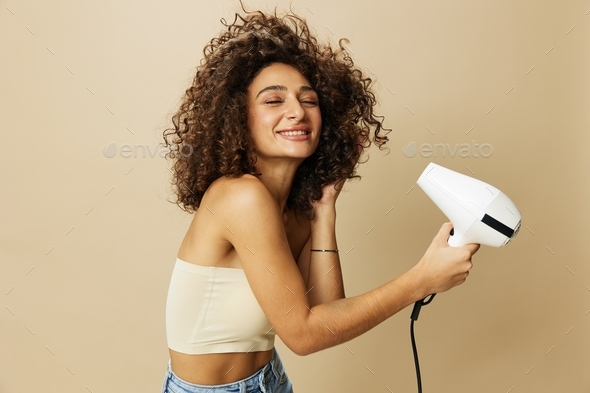 Woman dries curly afro hair with blow dryer, home beauty care styling products hair, smile on beige