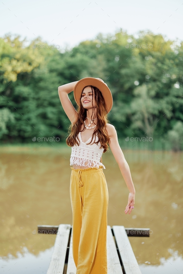 A young woman in a hippie look and eco-dress travels in nature by the lake wearing a hat and yellow