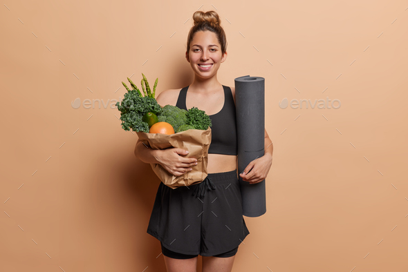 Sporty woman poses with bag of green vegetables and rolled fitness mat dressed in black tracksuit