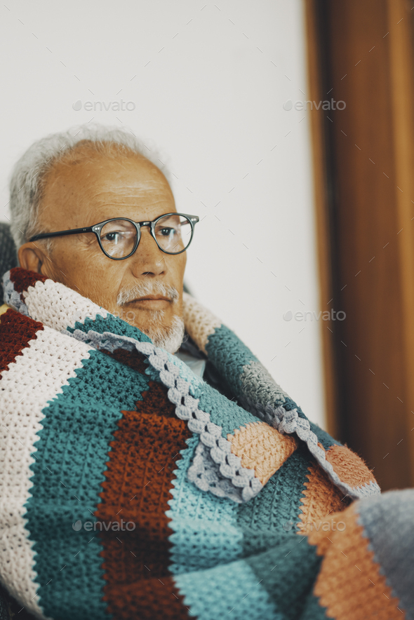 One old senior man sitting on the sofa at home with colorful wool cover for low temperature