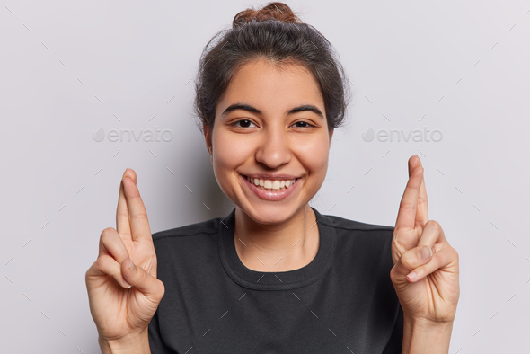 Positive Iranian girl with toothy smile keeps fingers crossed praying and hoping for good luck makes