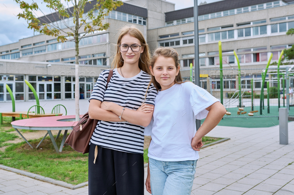 Portrait of two junior and high school schoolgirl sisters posing near school building