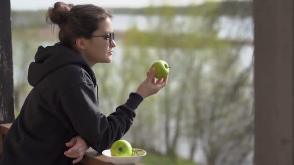 A Young Woman Holds an Apple in Her Hands and Enjoys Its Aroma Standing on the Veranda