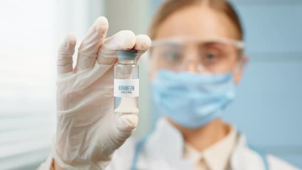 Blurred young woman medical nurse in disposable mask and glasses holds coronavirus vaccine alt